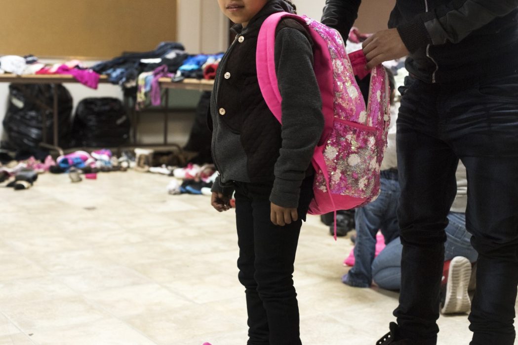 Small child in pink backpack stands in a room full of donated clothing while an adult puts something in her backpack.